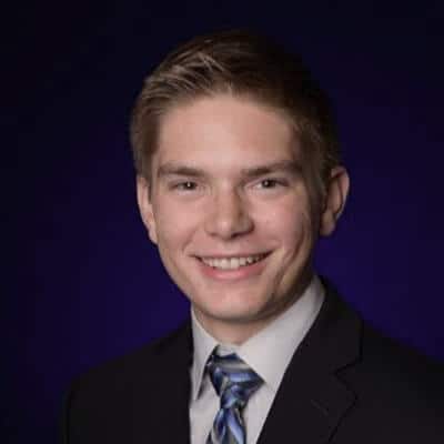 A smiling person in a dark suit and striped tie stands against a deep purple background, capturing the confident presence of Aaron Shishilla.