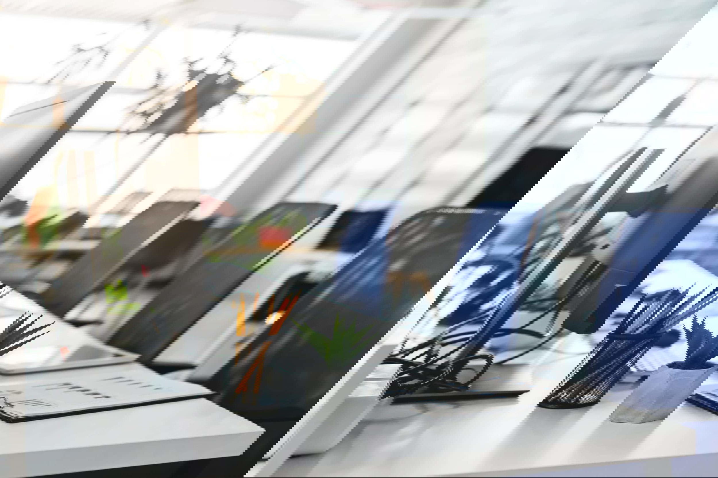 A modern office space featuring desks with blue chairs, perfect for freelancers comparing Upwork vs Fiverr. The desks boast monitors, a potted succulent, a cup of pencils, and a document folder. The office exudes a bright, airy vibe with plants and large windows in the background.