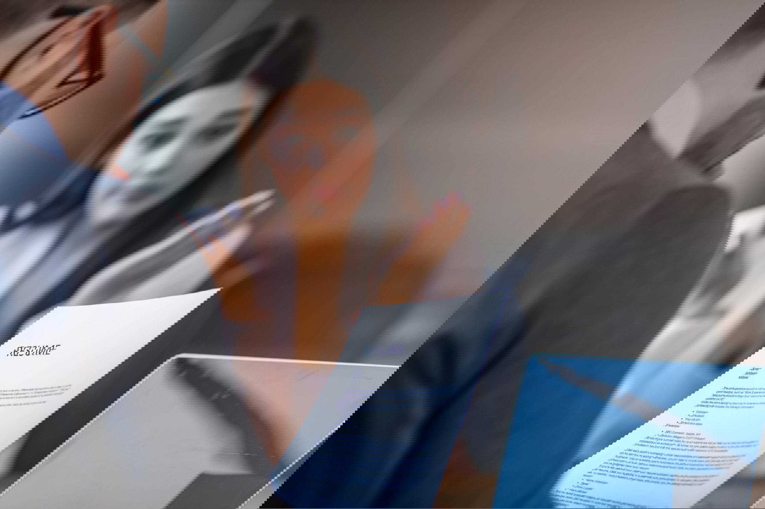 A person in business attire holds a resume while engaging with a woman in an office setting. The woman, possibly a virtual assistant, gestures animatedly, with an open laptop on the table. The focus is on the resume between them.