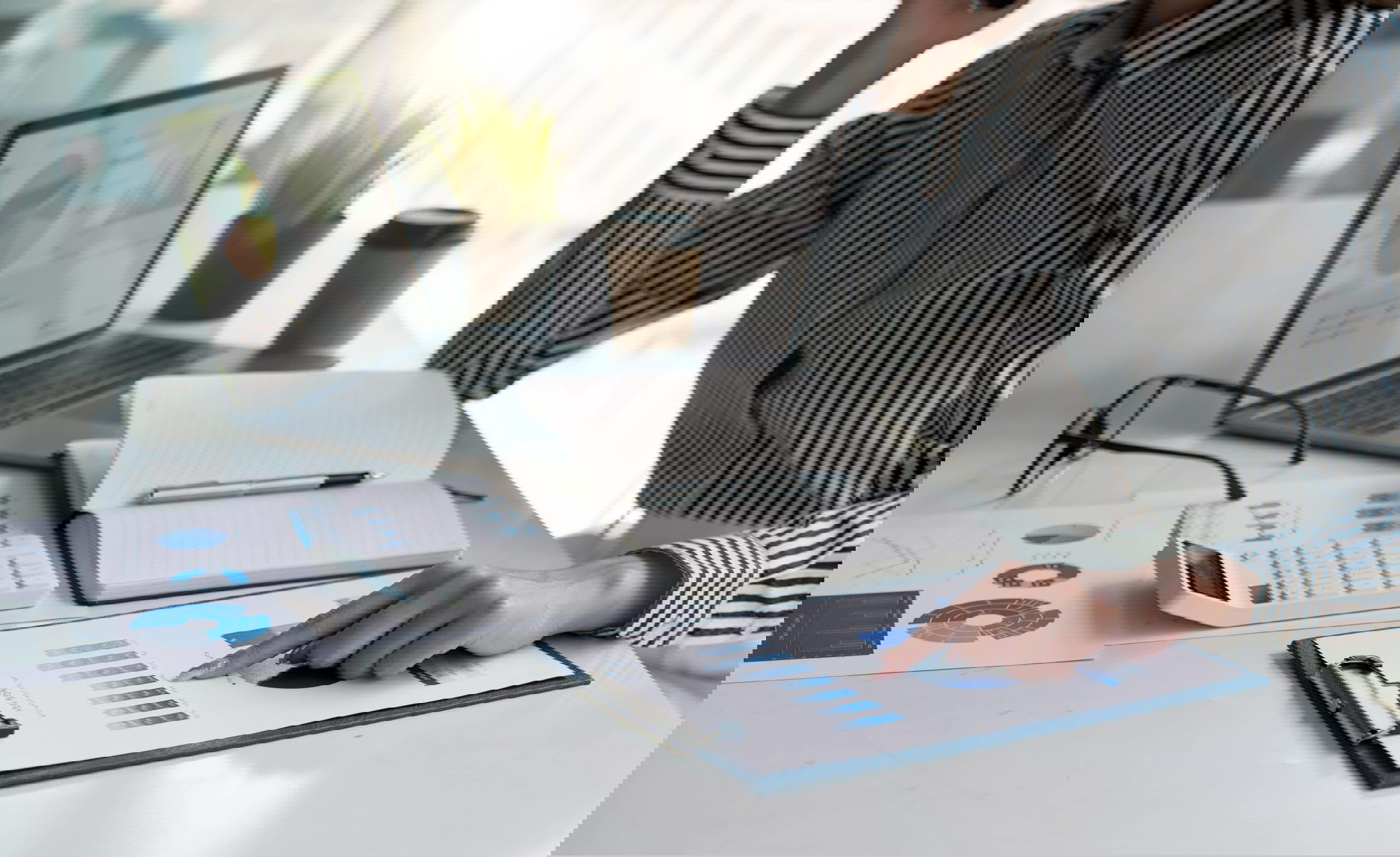 A person in a striped shirt reviews a graph on a clipboard at a desk, pondering the difference between a freelancer and a Virtual Assistant. Nearby, a laptop displays charts alongside an open notebook, calculator, eyeglasses, potted plant, and coffee cup as sunlight filters through the window.