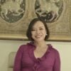 Jeanette Patindol, with short brown hair, is smiling and wearing a pink sweater with ruffled edges. She sits in front of an ornate tapestry embroidered with horses.