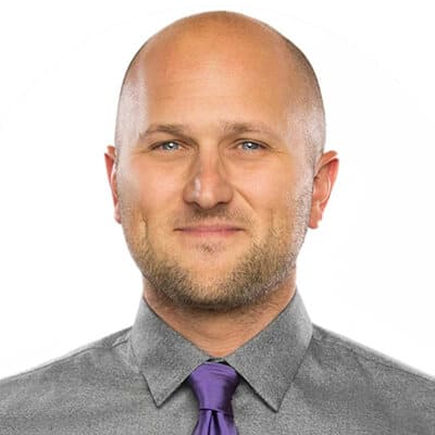 A bald man with a beard, wearing a grey shirt and a purple tie, smiles gently against a plain white background.