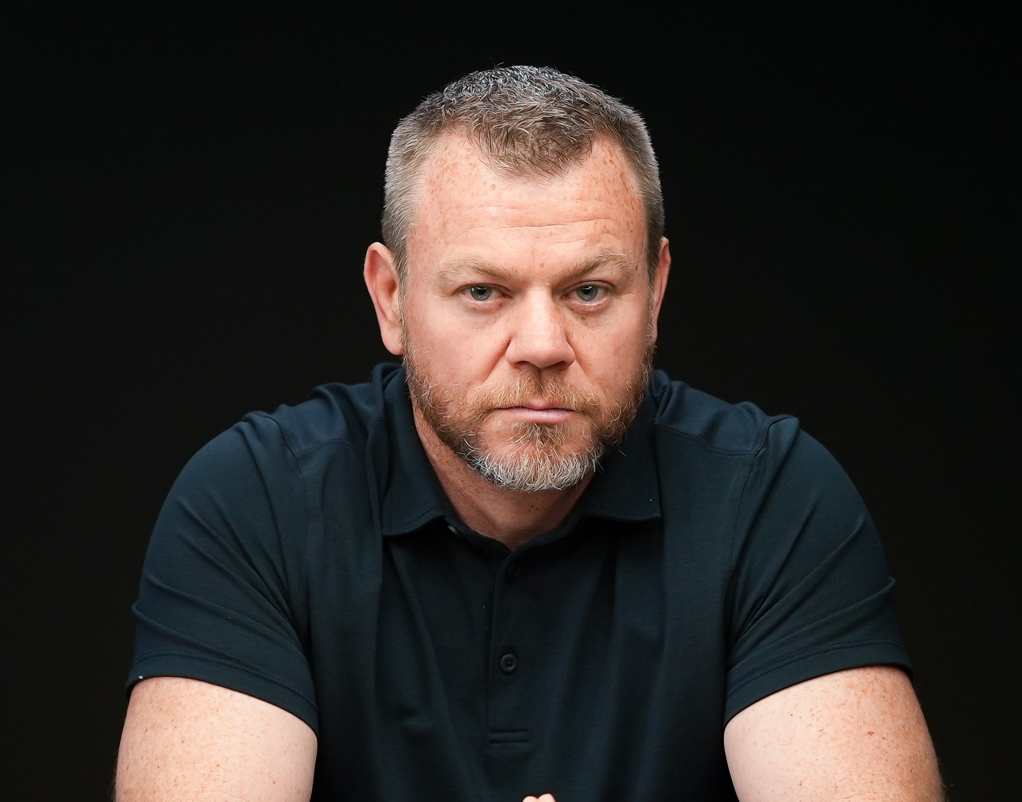 A man with short hair and a beard sits against a dark background, wearing a short-sleeved, dark polo shirt. He has a neutral expression and is looking directly at the camera, embodying the professional demeanor often associated with MyOutDesk.