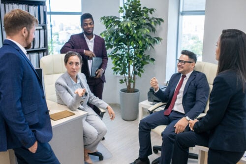 Five colleagues in a modern office engage in discussion. Two are seated, and three stand near desks with paperwork and computers, likely discussing how to delegate tasks efficiently. A potted plant is visible by the window. Everyone wears business attire, underscoring the professional atmosphere.