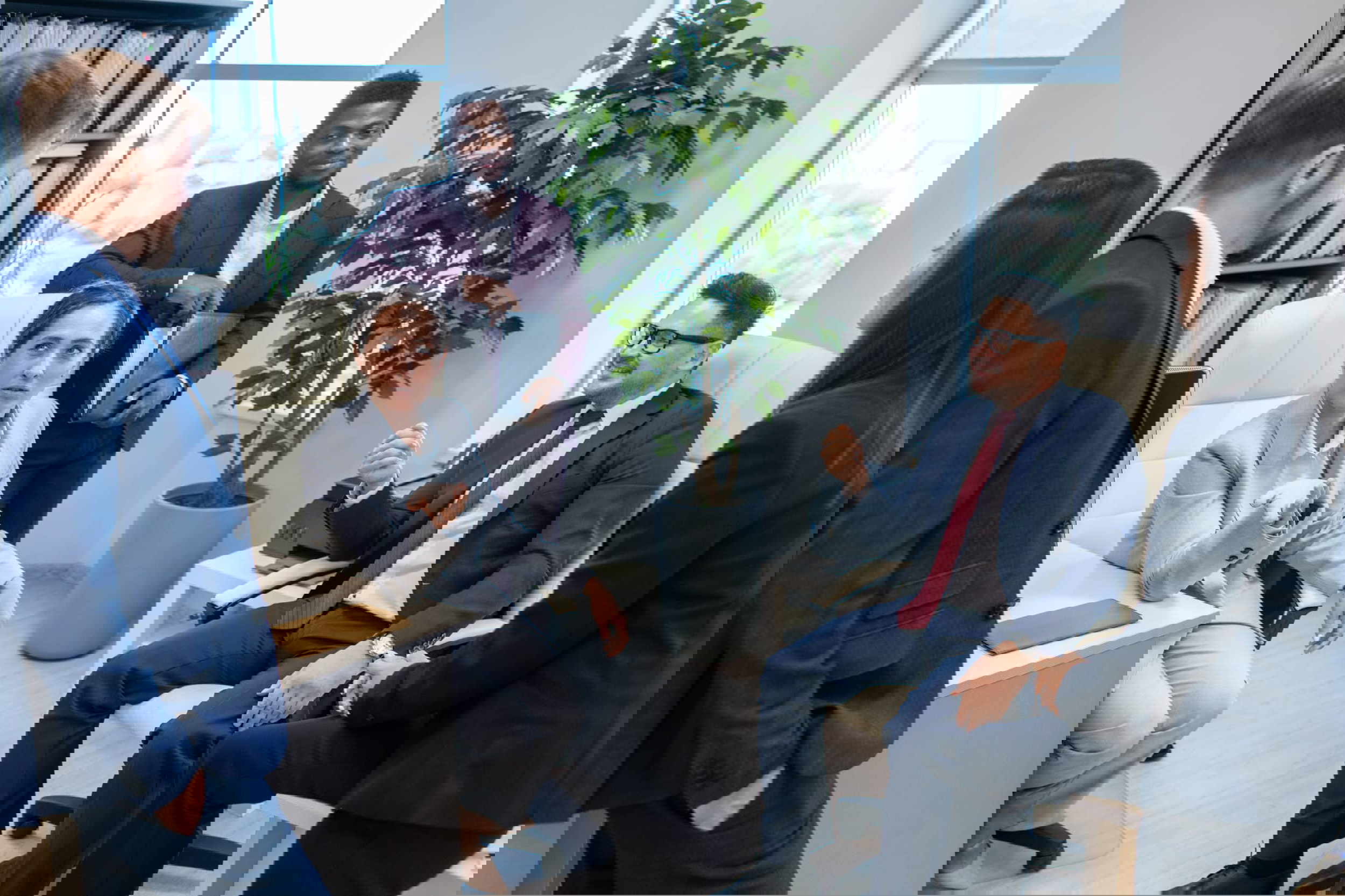 Five colleagues in a modern office engage in discussion. Two are seated, and three stand near desks with paperwork and computers, likely discussing how to delegate tasks efficiently. A potted plant is visible by the window. Everyone wears business attire, underscoring the professional atmosphere.