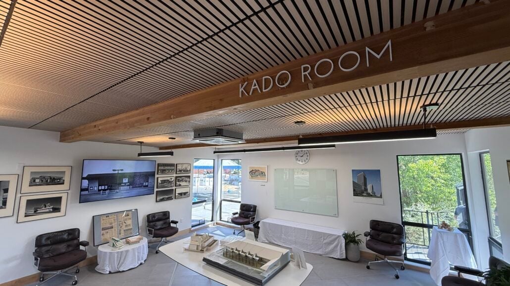 A modern meeting room named KADO ROOM at the MyOutDesk Sacramento headquarters grand opening features wooden ceiling beams, chairs, framed photos on the walls, a model building on a central table, and large windows letting in natural light.