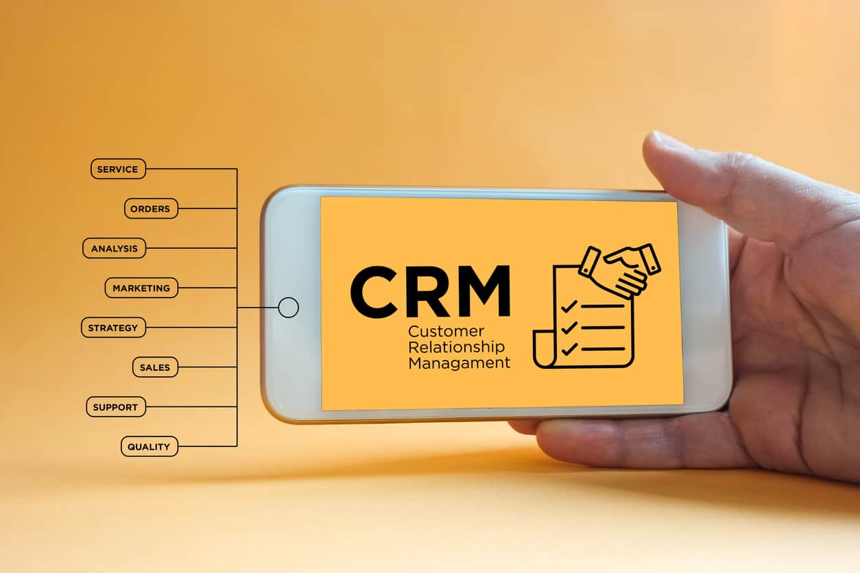 A hand holds a smartphone displaying CRM Customer Relationship Management with an icon of shaking hands and a checklist. To the left, there are connected labels: service, orders, analysis, marketing, strategy, sales, support, quality.