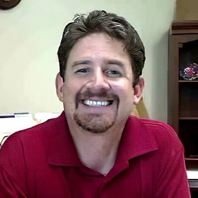 Jeff Robbins, with short dark hair and a goatee, smiles at the camera. He wears a red collared shirt while sitting indoors, surrounded by a backdrop of bookshelves and scattered papers.