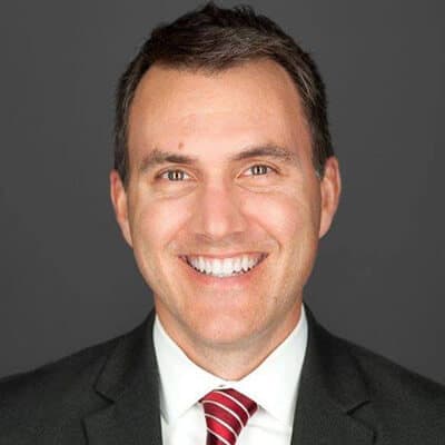 Smiling person in a suit and striped tie against a gray background.