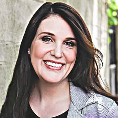 Keeley Teemsma, a smiling woman with long dark hair, stands against a textured wall. She wears a gray blazer over a black top, with lush greenery in the background adding a touch of nature to her poised and confident presence.