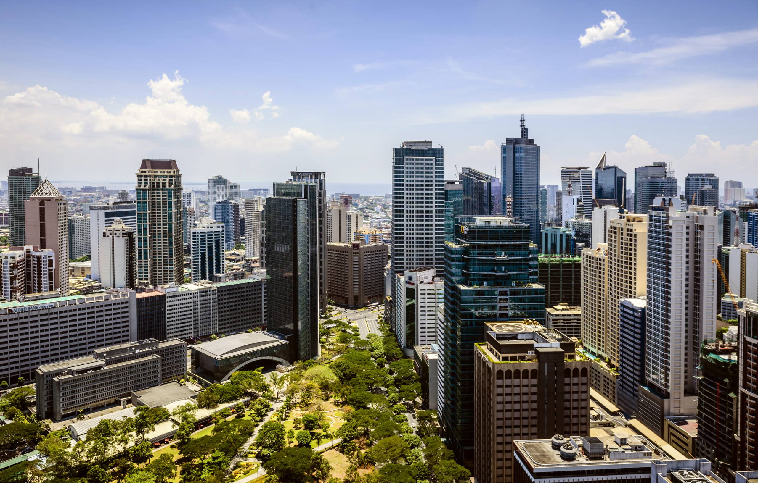 Aerial view of a modern city skyline with numerous skyscrapers and high-rise buildings. The foreground features a green park with trees. The Philippines, known for being a top destination where major corporations outsource their business needs, boasts clear skies dotted with clouds.