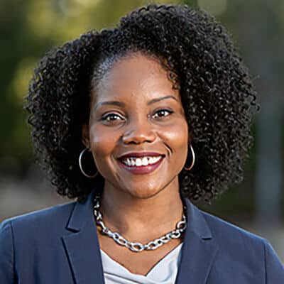 Smiling person with curly hair, identified as Shawana Hudson, wearing a navy blazer, hoop earrings, and a silver chain necklace. The background is a beautifully blurred outdoor setting.