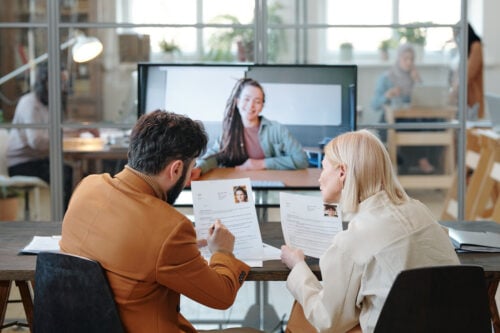 Two people sit at a table with resumes, conducting a video interview in the bright, modern office. The candidate appears on a large screen in this guide on how to hire virtual employees and why you should. Through the glass wall, more colleagues are visible working diligently in the background.