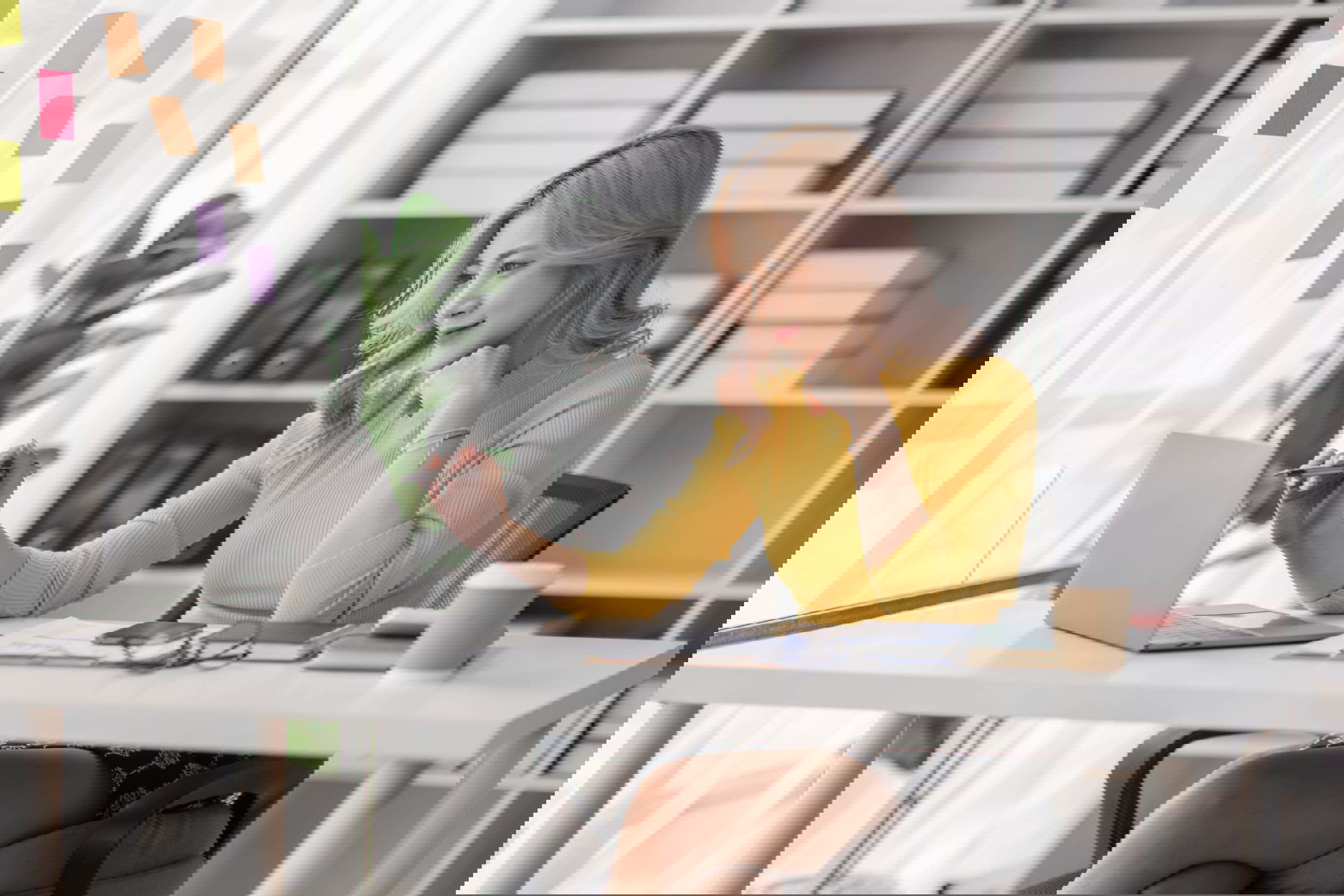 A person with blonde hair in a yellow sweater sits at a desk, engrossed in researching Zirtual alternatives on their laptop. The desk is cluttered with papers, glasses, a pen, and a coffee cup. Shelves filled with boxes and binders line the background, while colorful sticky notes adorn the window.