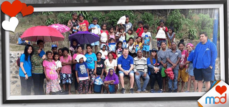 A group of people, including adults and children, stand and sit outdoors in front of greenery, celebrating the joy of giving. Some hold umbrellas. The image is framed with heart decorations and a logo in the corner, capturing the essence of this heartfelt gift moment.