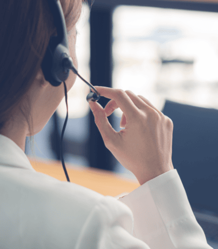 A person wearing a white shirt is seen from behind, using a headset with a microphone. They appear to be in an office setting, engaging in a call or teleconference. The background is softly blurred with a hint of window light.